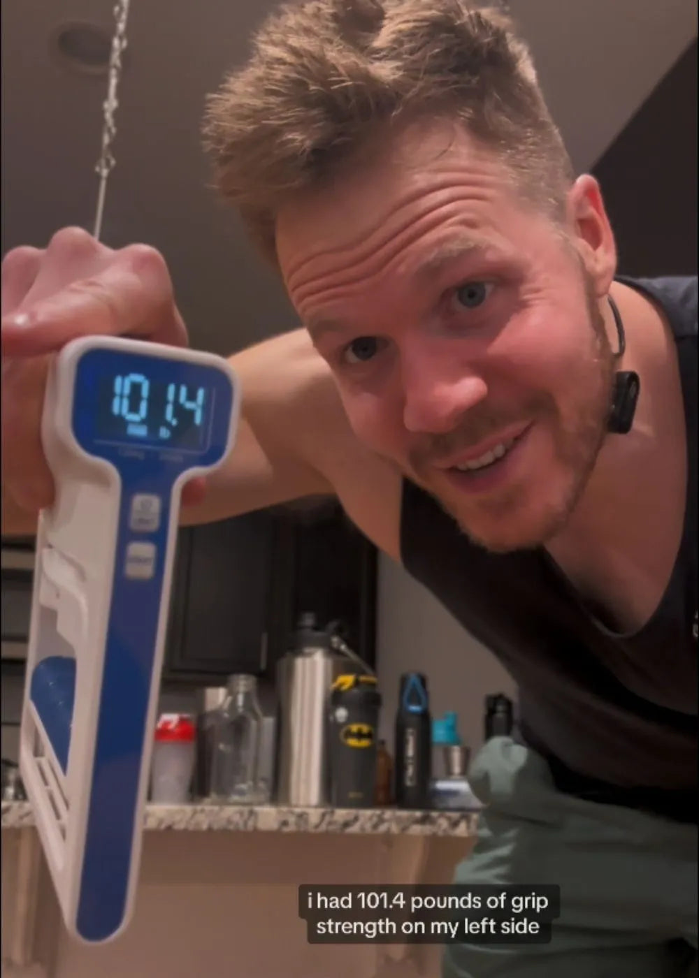 A person holding a digital grip strength hand dynamometer. The device displays "101.4" on its screen, indicating grip strength measurement. The background shows a kitchen setting with various items. Keywords: grip strength hand dynamometer, digital grip strength tester, grip strength measurement tool.