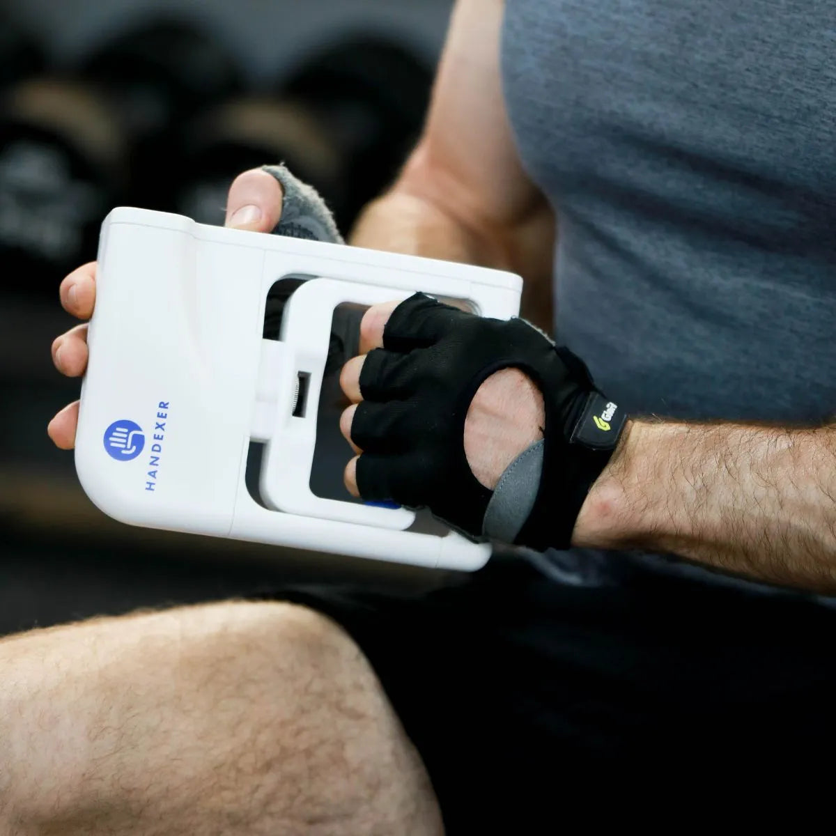 A person wearing black fingerless gloves uses a Handexer grip strength hand dynamometer, a tool for measuring grip strength, with the Handexer logo clearly visible on the white device.