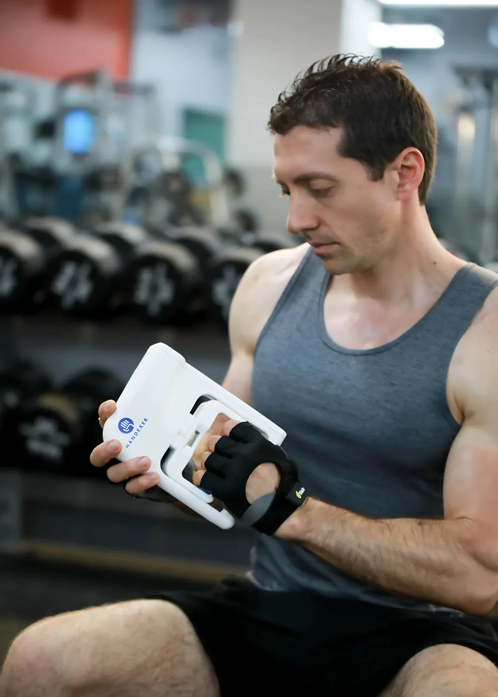 A man in a gym setting, wearing a gray tank top and black shorts, is using a Handexer white grip strength hand dynamometer. He has black workout gloves on his right hand, which is gripping the dynamometer. In the background, there are rows of dumbbells. This image showcases a Handexer grip strength measurement tool, ideal for isometric grip exercises and testing hand grip strength. Keywords: Handexer, grip strength hand dynamometer, grip strength measurement tool, isometric grip exercises