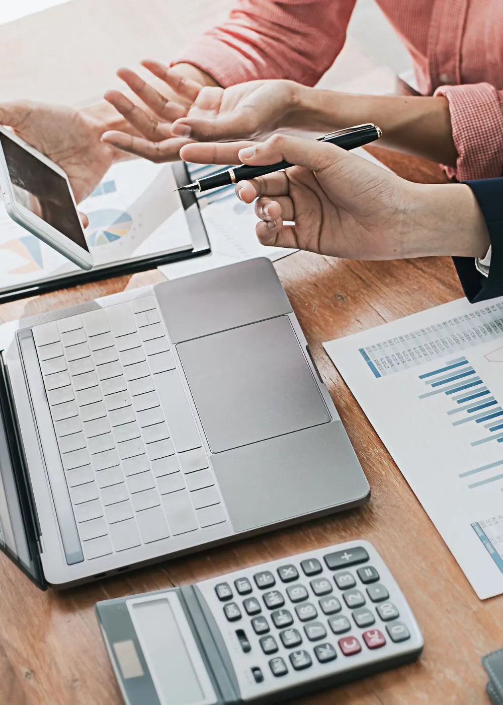 Two hands interacting over a workspace with a Handexer grip strength hand dynamometer - related items like a laptop, calculator, and documents. One hand holds a pen, indicating a professional setting for grip strength measurement tool discussions.
