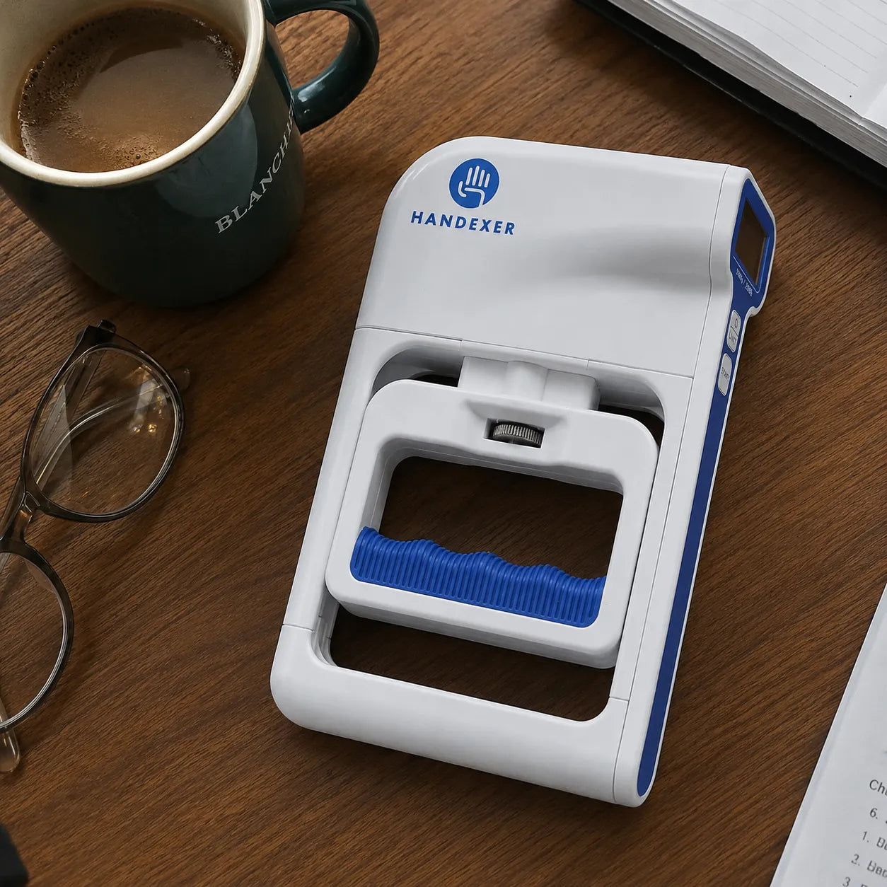 Handexer digital hand dynamometer on a wooden desk beside coffee, glasses, and an open notebook