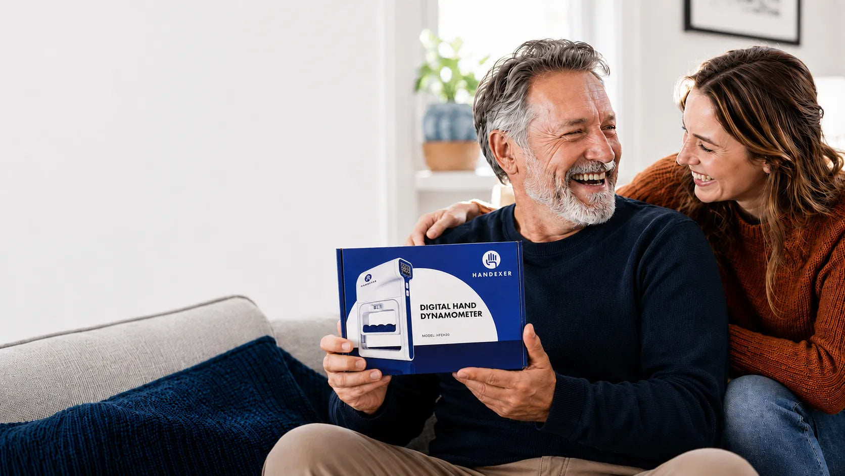 Smiling man and woman holding a Handexer digital hand dynamometer box on a sofa in a bright home setting