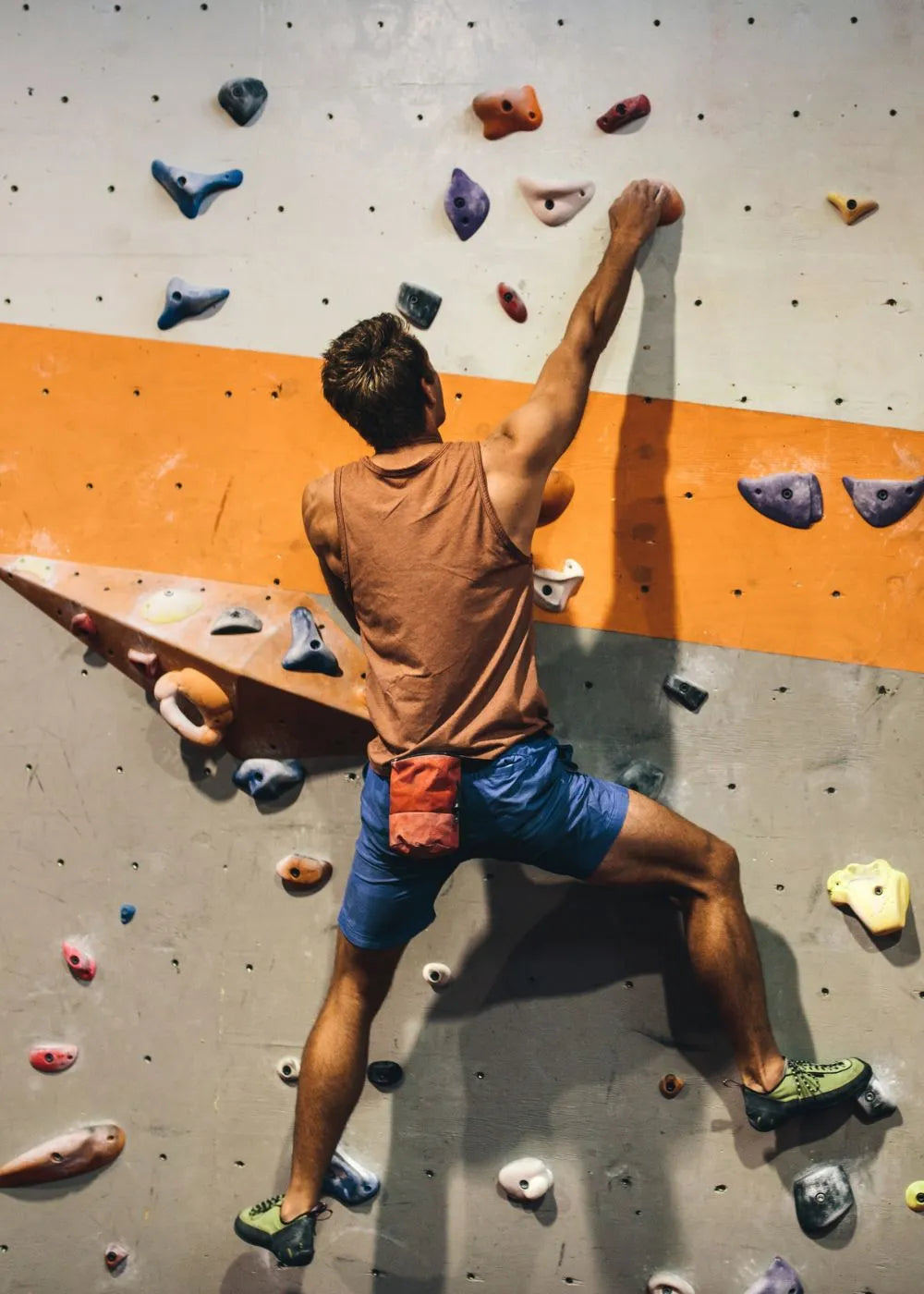 A person wearing a brown tank top and blue shorts is climbing an indoor rock wall with colorful holds. The climber is using their hand grip strength, which is essential for such activities, and this image relates to grip strength measurement tools like the Grip Strength Hand Dynamometer as it showcases the importance of hand grip muscles in climbing.