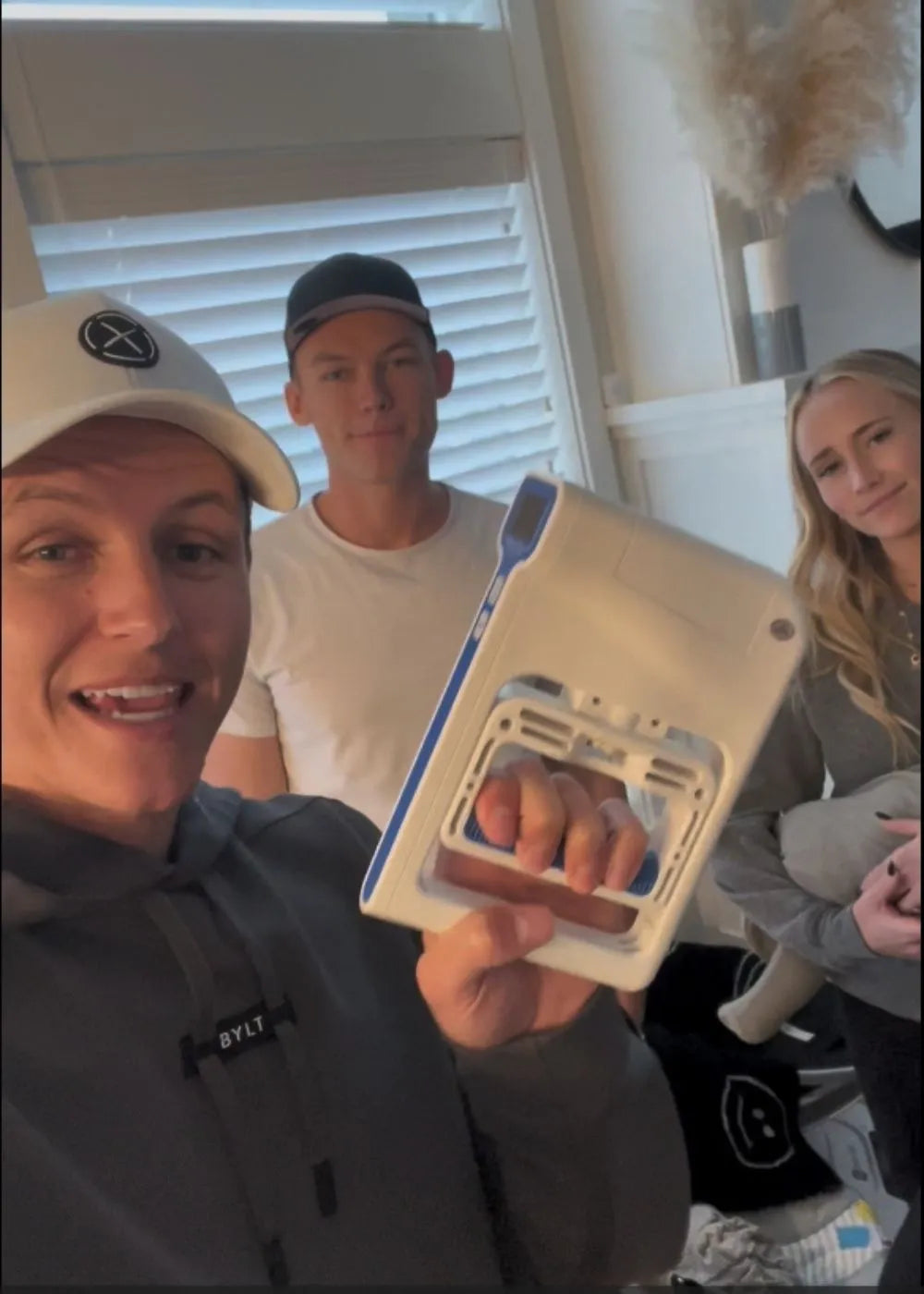 A person wearing a gray hoodie and a white cap is holding a white and blue grip strength hand dynamometer. Two other individuals are standing in the background. The setting appears to be indoors with a window and some decor visible. Keywords: grip strength hand dynamometer, grip strength measurement tool, hand dynamometer