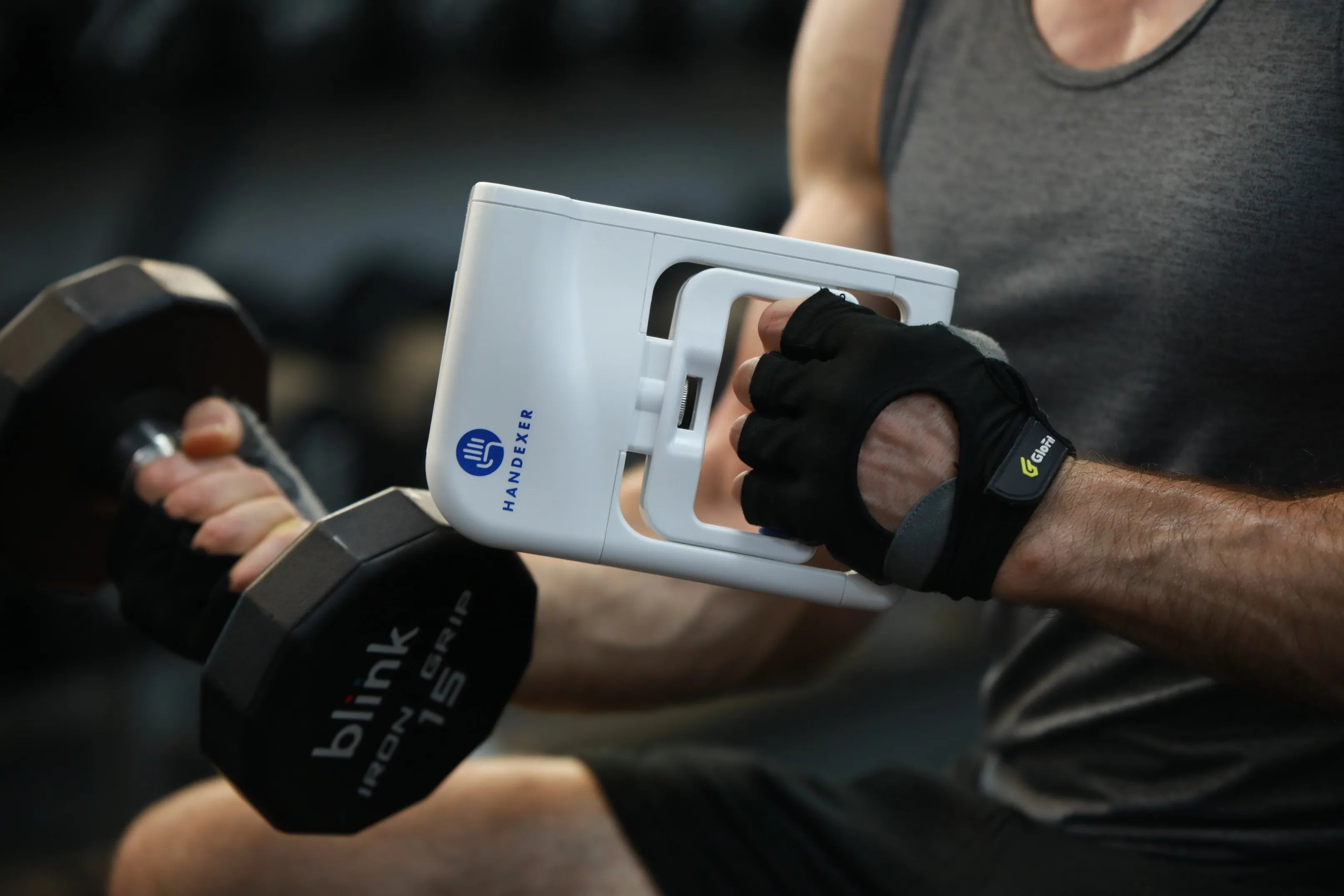 A person wearing black workout gloves is using a white HandExer grip strength hand dynamometer. The device is being held in one hand while the other hand grips a black dumbbell labeled "blink IRON GRIP 15RIP." The HandExer device has a blue logo and text on it, indicating it is a tool for grip strength measurement. This setup is ideal for isometric grip exercises and training hand grip muscles. Keywords: HandExer, grip strength hand dynamometer, grip strength measurement tool, isometric grip exercises, hand