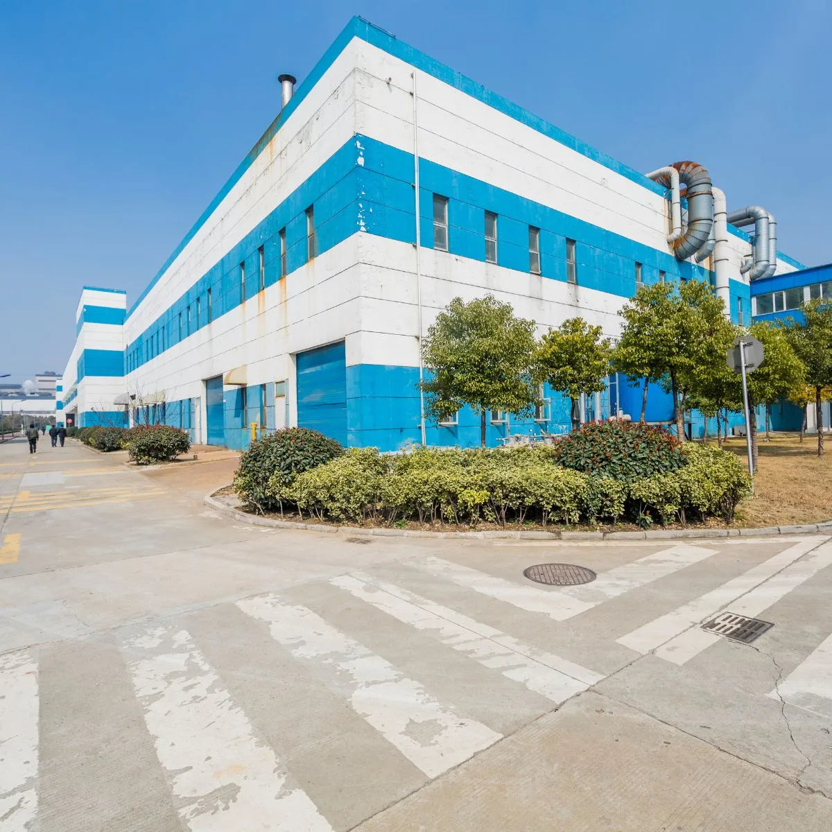 A blue and white industrial building, likely a factory, with visible pipes and ventilation systems. The building is surrounded by greenery, including trees and bushes, and there is a paved area in front. This could be the manufacturing facility for Handexer's grip strength hand dynamometers and other grip strength measurement tools.