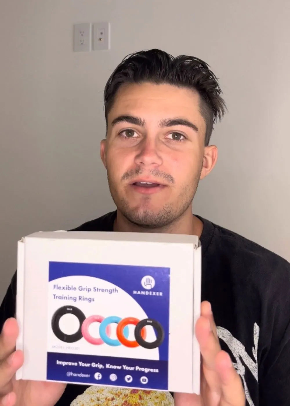 A person holding a box of "Flexible Grip Strength Training Rings" by Handexer. The box features images of colorful grip rings and text promoting grip improvement and progress tracking. Keywords: grip strength, training rings, Handexer, grip improvement.