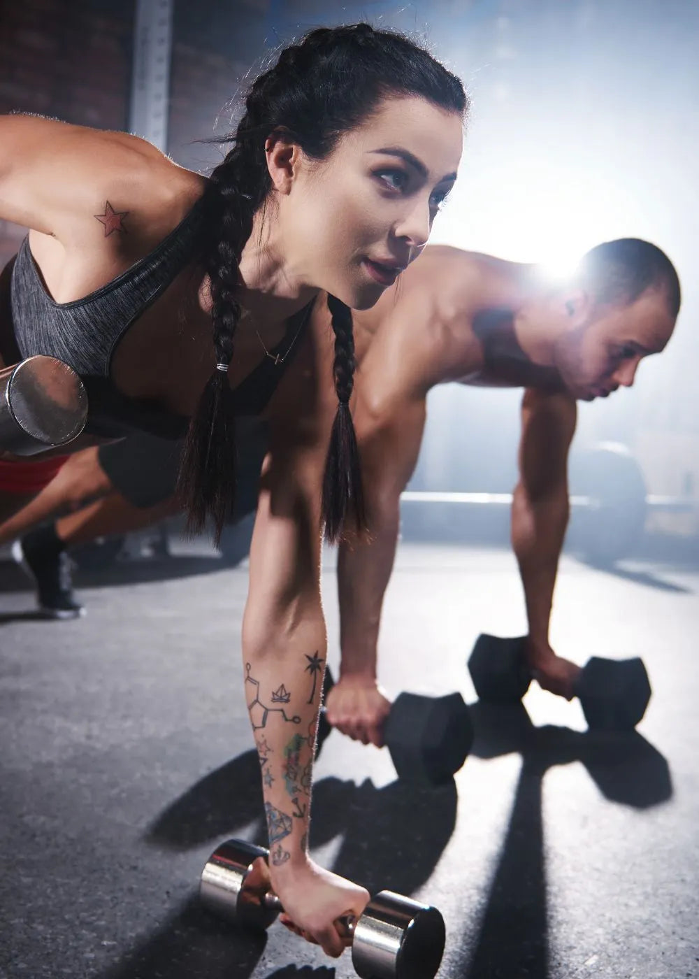 Two individuals performing plank exercises with dumbbells in a gym setting. One person has braided hair and visible tattoos on their arm. This image relates to grip strength training, as grip strength is crucial for holding dumbbells during such exercises. Keywords: grip strength measurement tool, isometric grip exercises, Handexer, grip strength tester.
