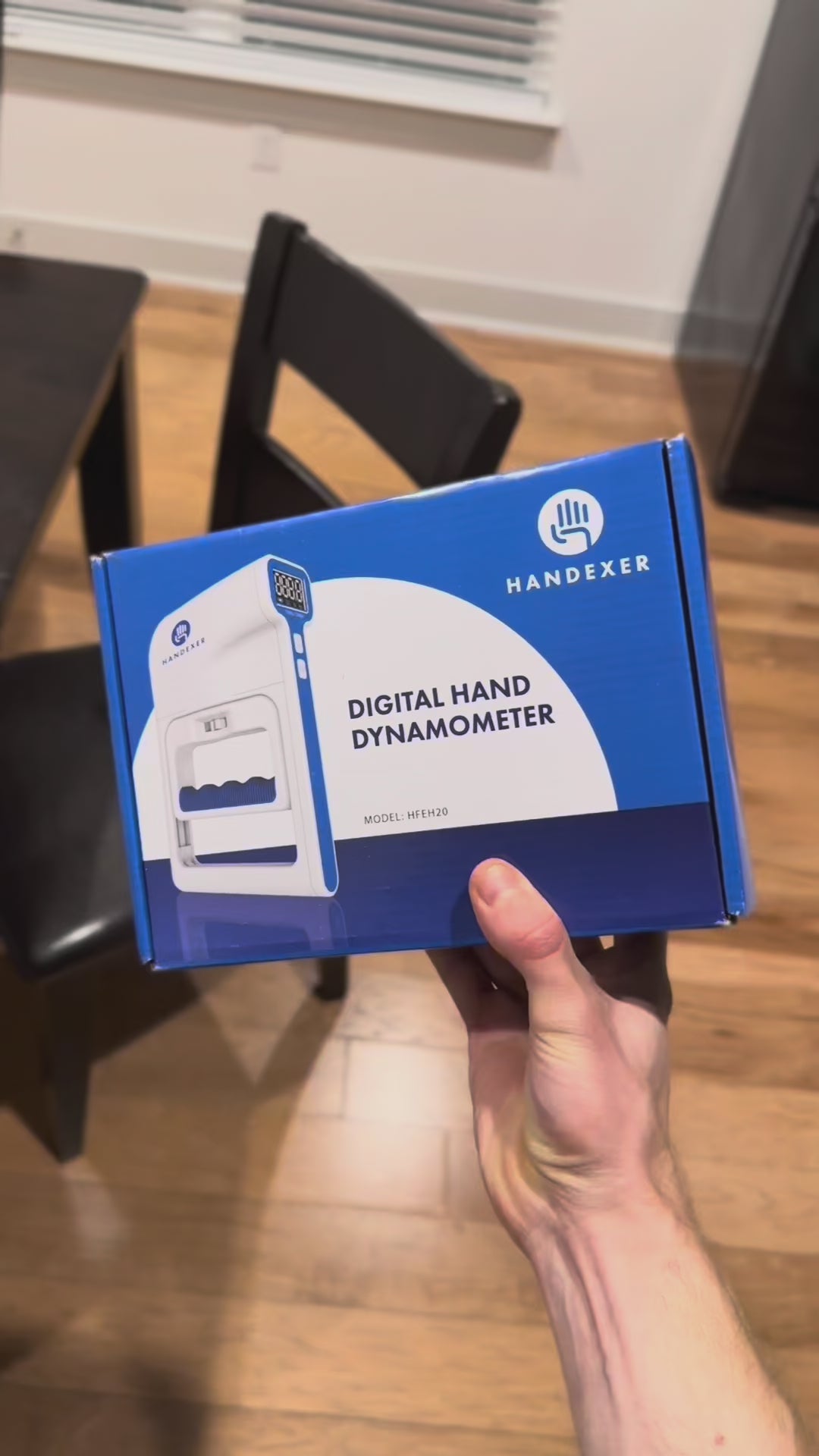 Load video: A person holding a digital grip strength hand dynamometer. The device displays "101.4" on its screen, indicating grip strength measurement. The background shows a kitchen setting with various items. Keywords: grip strength hand dynamometer, digital grip strength tester, grip strength measurement tool.